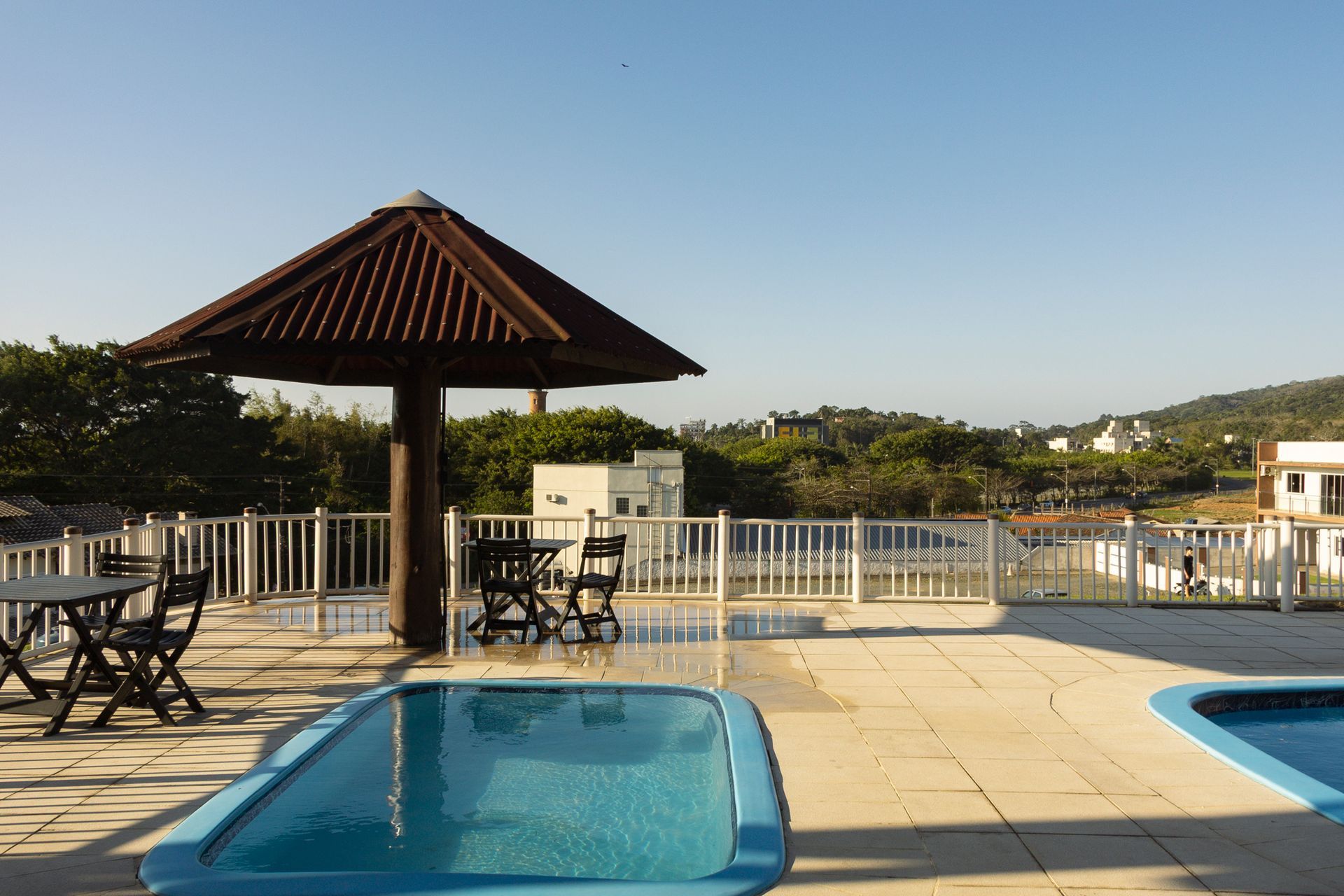 Área da piscina com guarda-sóis, mesas e vista para a vegetação e o céu.