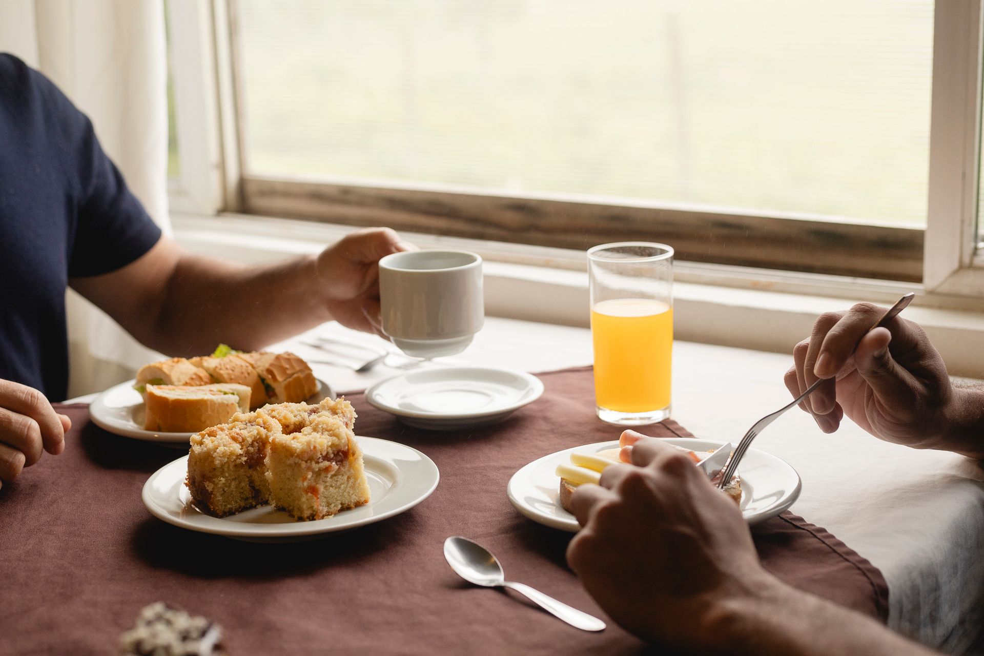 Duas pessoas tomando café da manhã perto de uma janela. Pratos com doces, suco de laranja e uma xícara de café sobre a mesa.