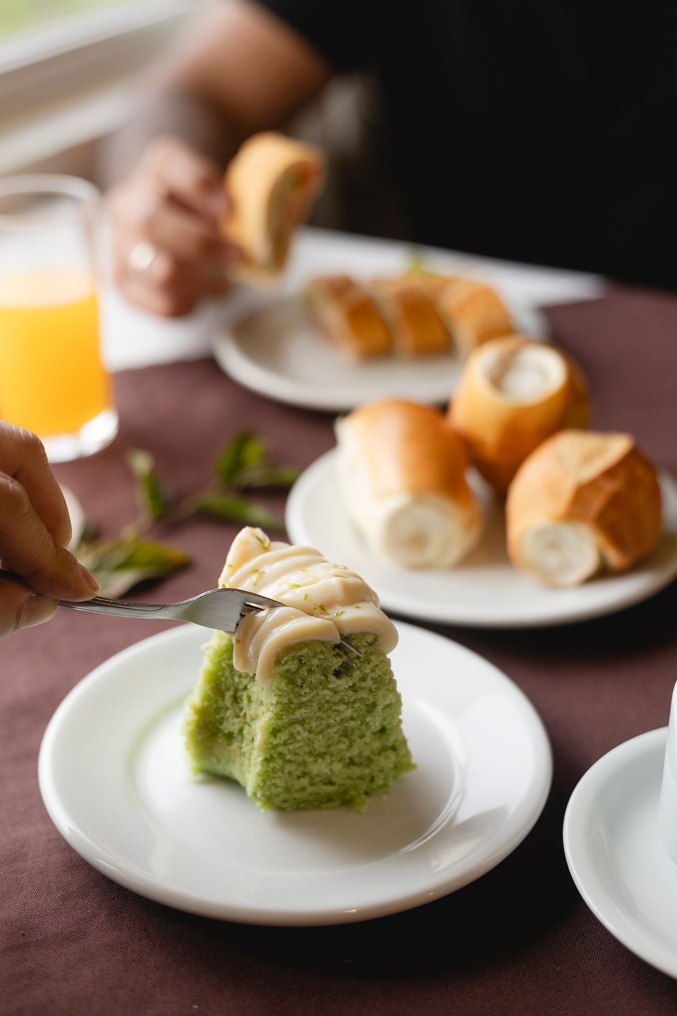 Bolo verde em um prato branco sendo cortado com uma faca; pão e suco de laranja sobre a mesa.