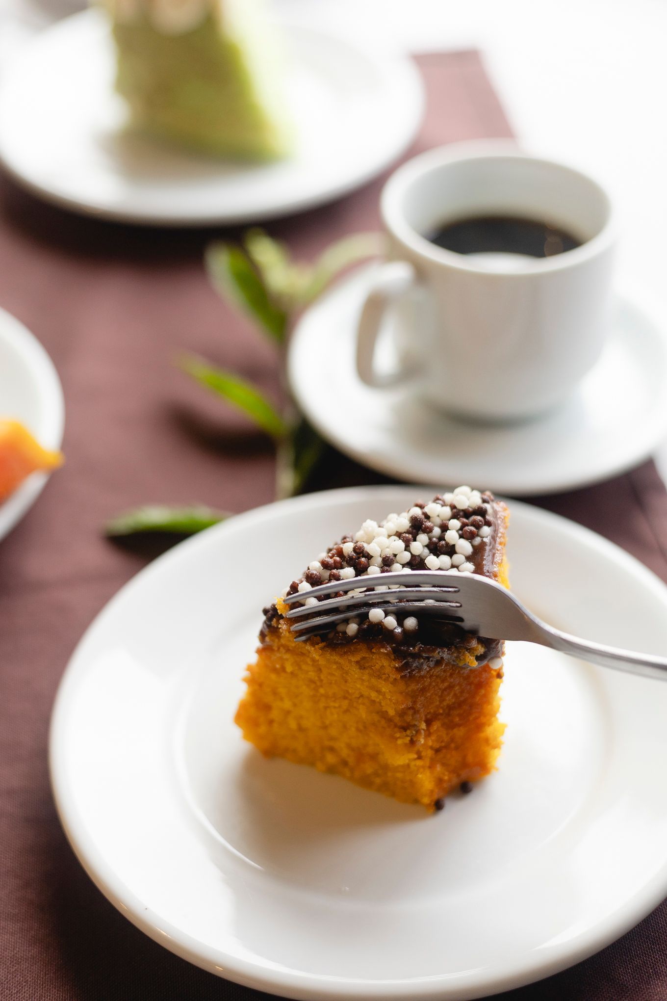 Fatia de bolo de cenoura com cobertura de chocolate, garfo, xícara de café e outra fatia de bolo sobre a mesa.