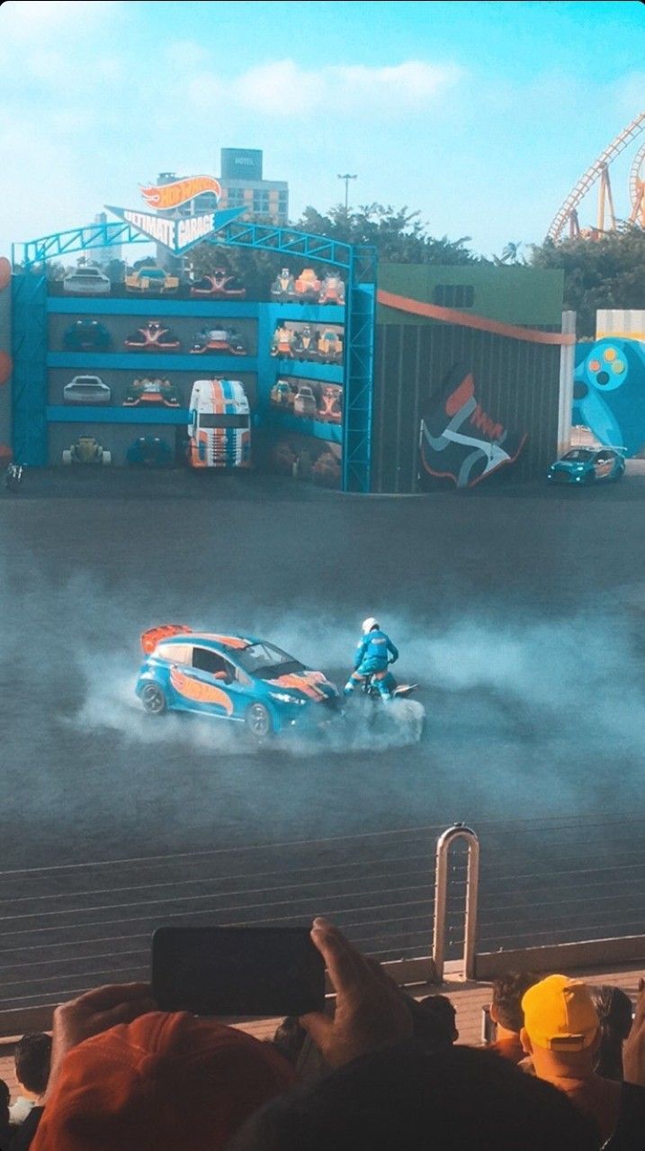 Um carro azul gira em um palco, com uma pessoa em cima. Ao fundo, uma exposição de carros e uma plateia.