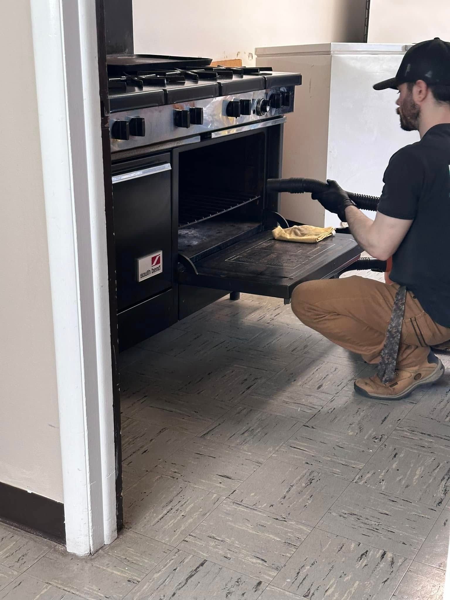 A person wearing gloves and a cap cleans an oven in a commercial kitchen.