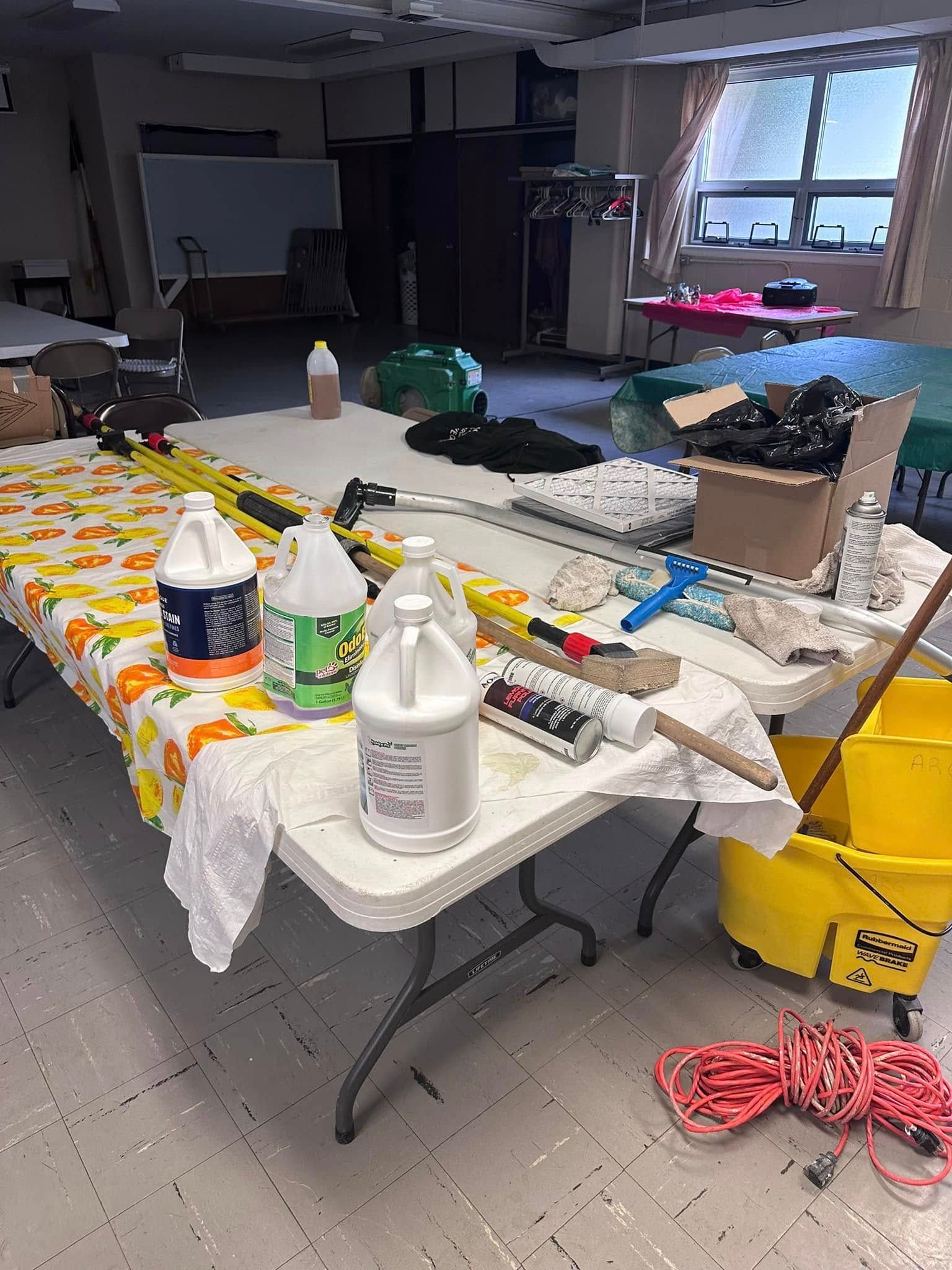 Cleaning supplies on tables in a room, including buckets, bottles, mops and a power cord.