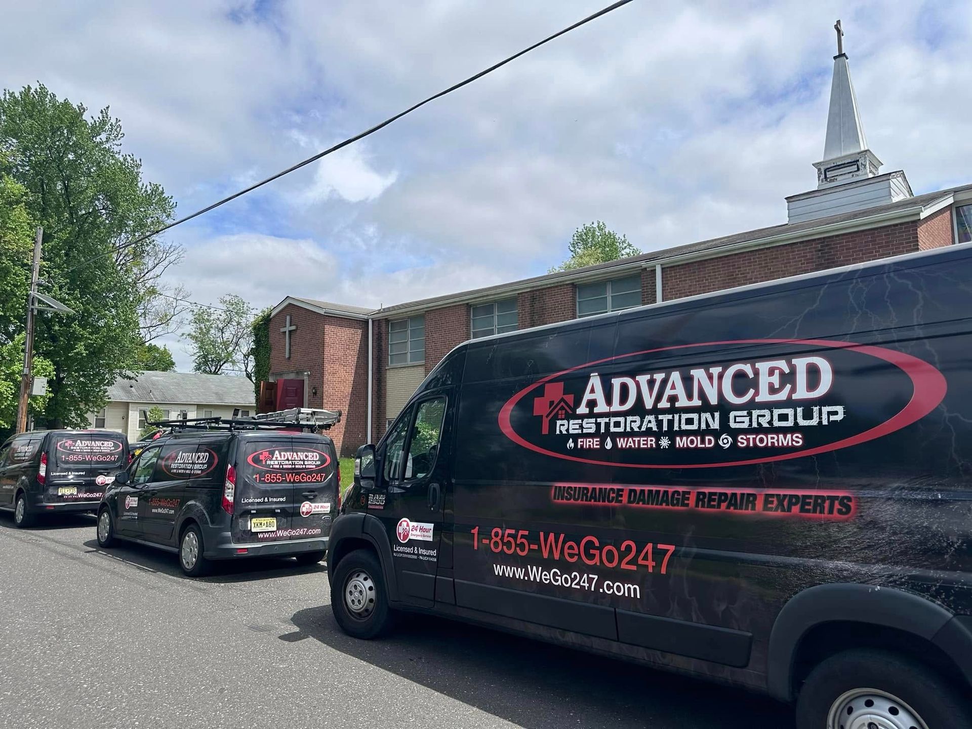 Three Advanced Restoration Group vans parked in front of a brick building and a church with a steeple.