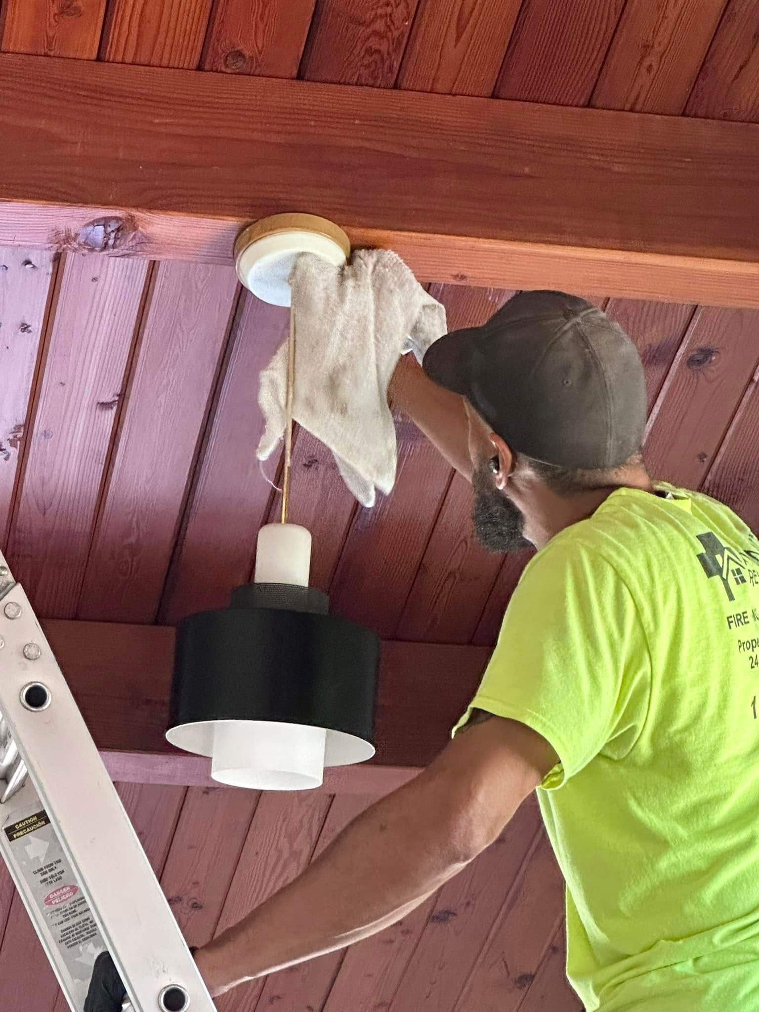 Person in a neon shirt installs a light fixture on a wooden ceiling, using a ladder.