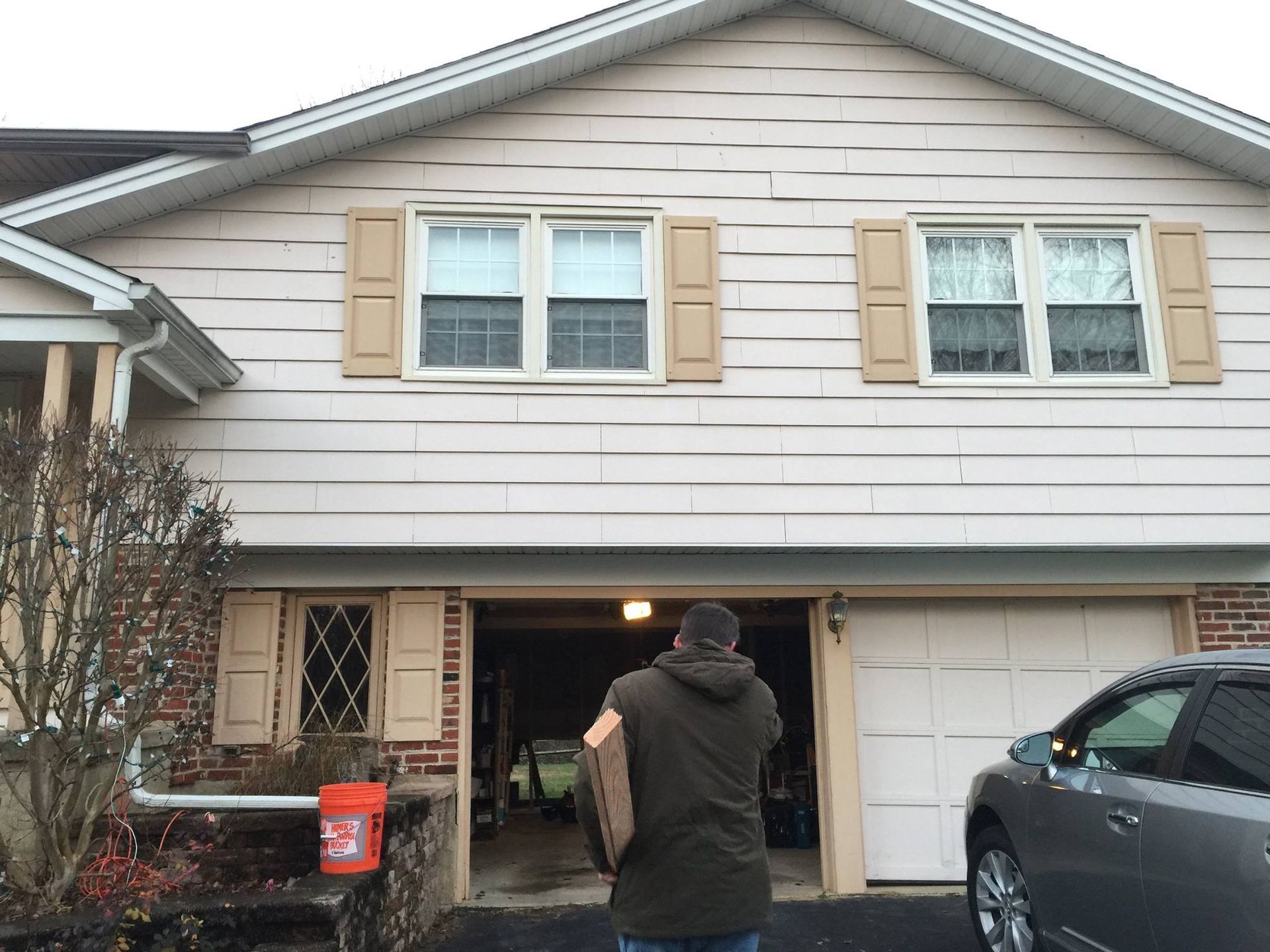 Man carrying item towards open garage door of a two-story house with beige siding and tan shutters.