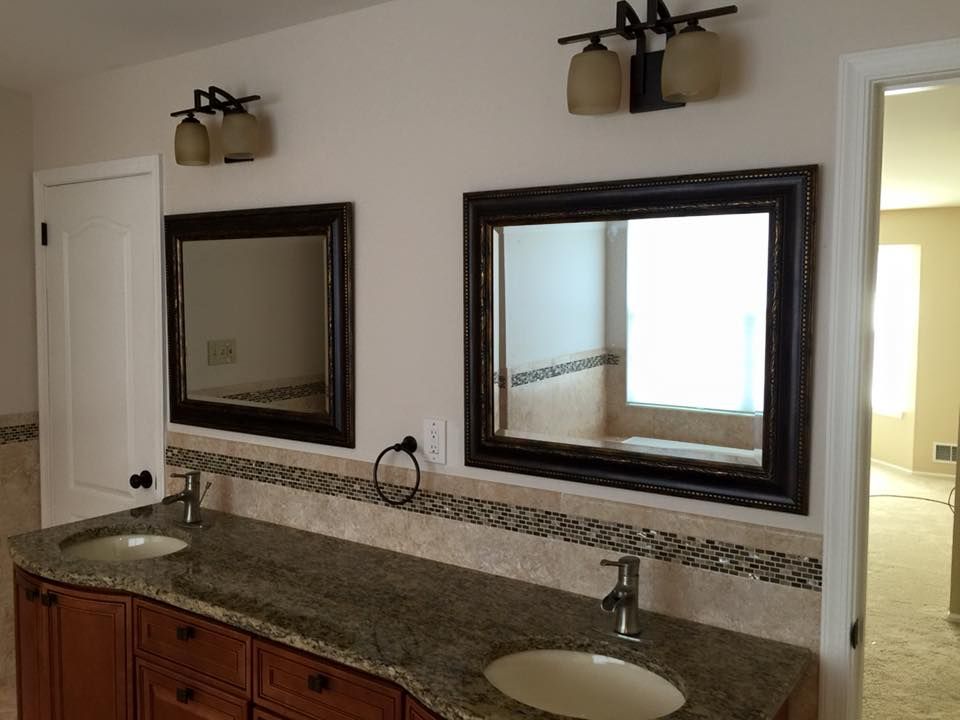 Bathroom with dual sinks, mirrors, and lighting fixtures. Brown cabinetry, granite countertop, and neutral walls.