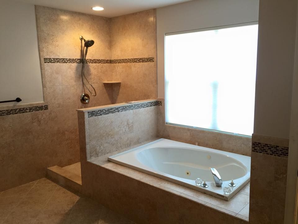 Bathroom with a built-in tub, beige tile, a showerhead, and a large window with frosted glass.
