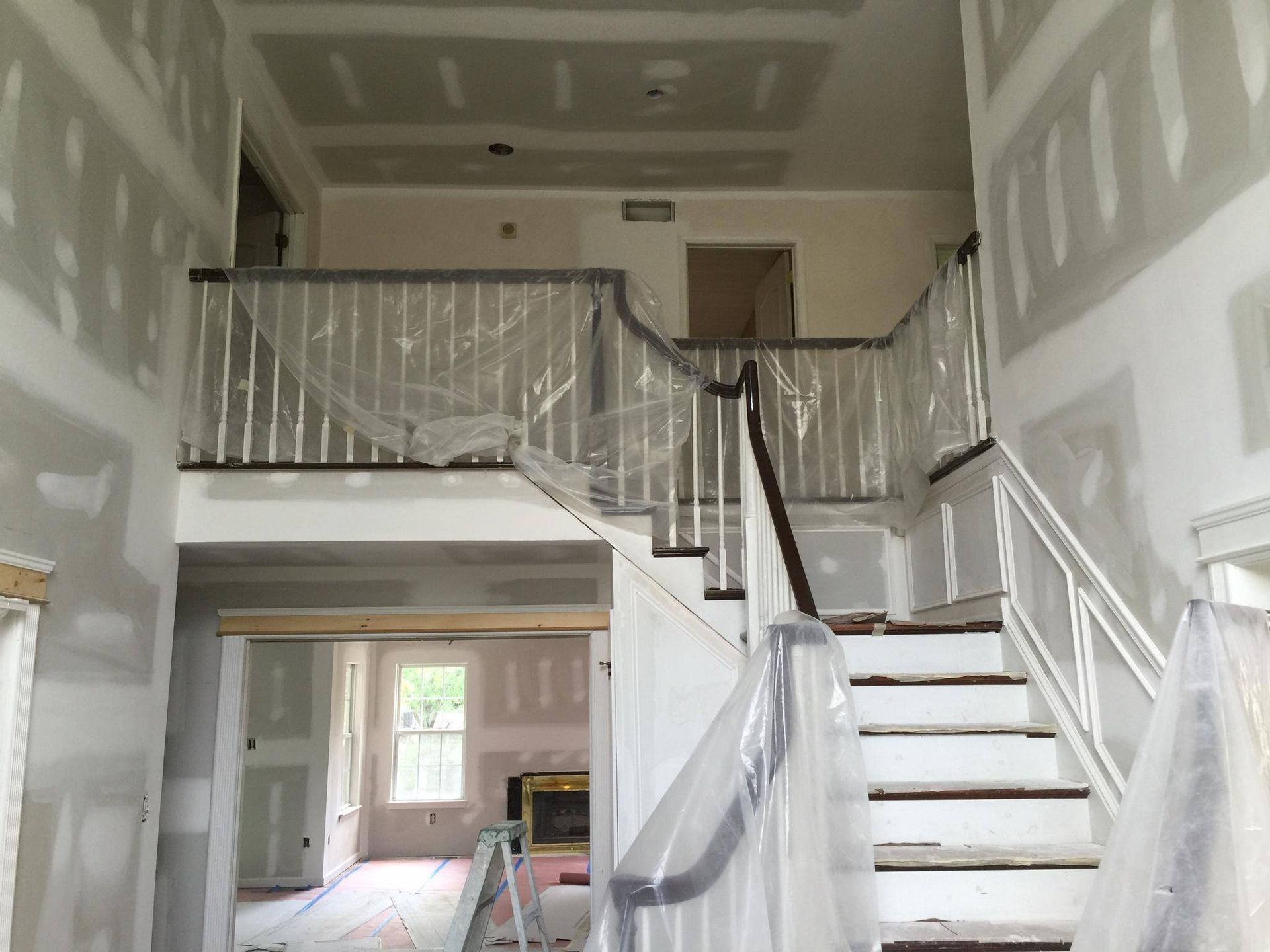 Interior of a house under construction; staircase, drywall, plastic wrap, and doorway visible.
