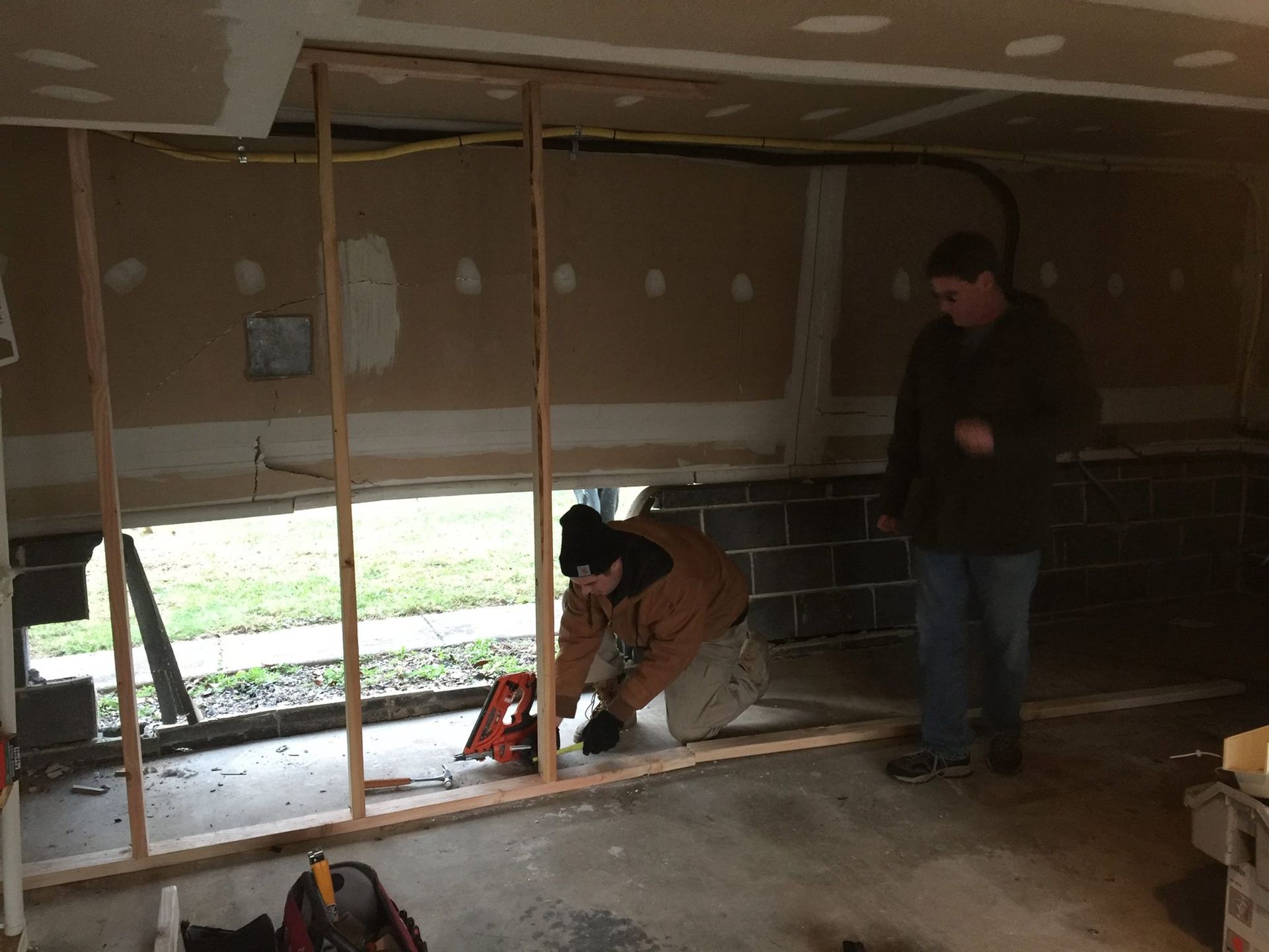 Two people building a wall in a garage. One cuts wood with a saw. The other watches.