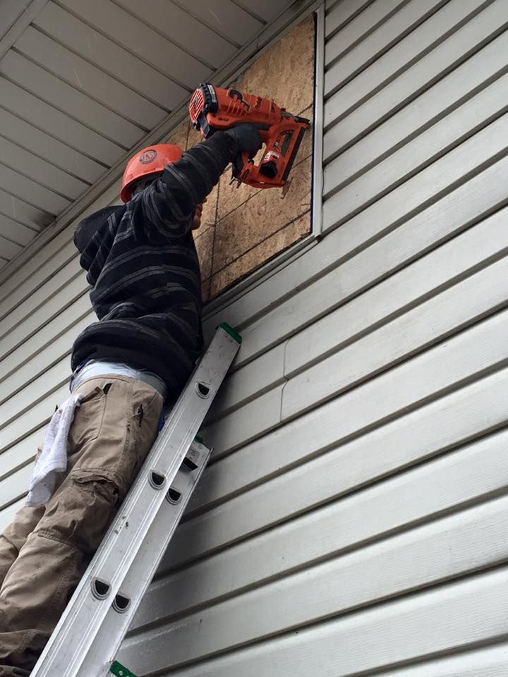 Person on ladder boards up a window with a nail gun on a building with white siding.