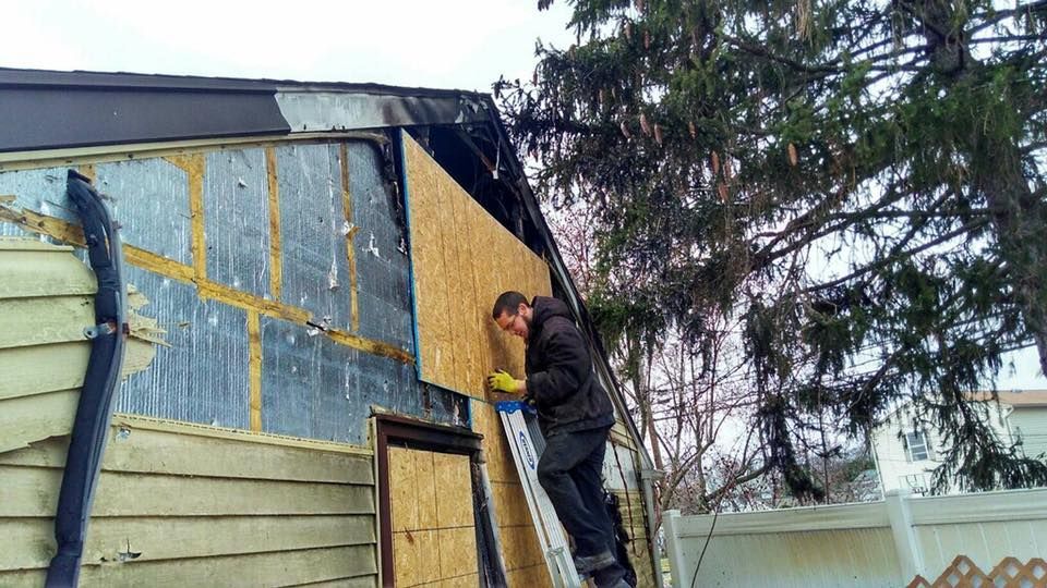 A person boards up a damaged building. They are on a ladder, wearing gloves, and working on the side of a house.