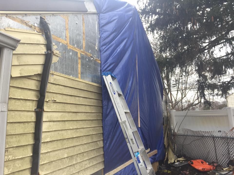 Damaged house exterior covered with blue tarp, ladder leans against it. Yellow siding and black downspout visible.