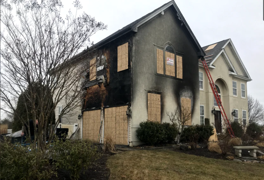 A two-story house with significant fire damage on the side, boarded-up windows, and a ladder against the other side.