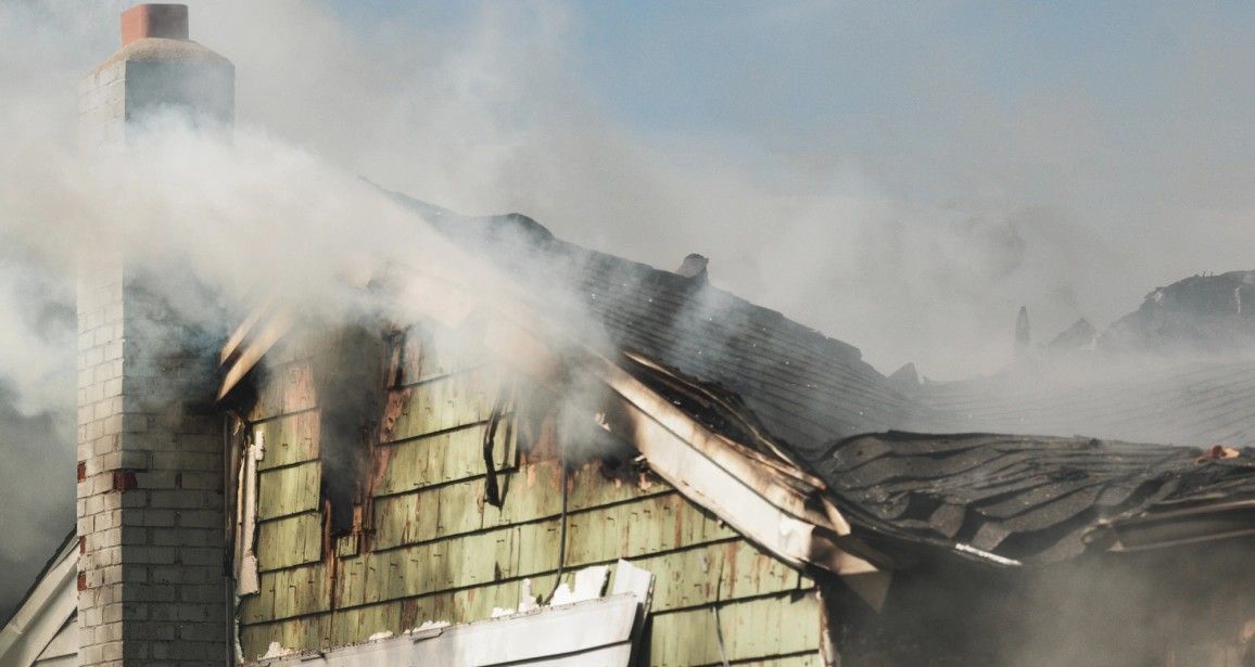 House roof on fire, billowing smoke, chimney visible, green siding, blue sky.