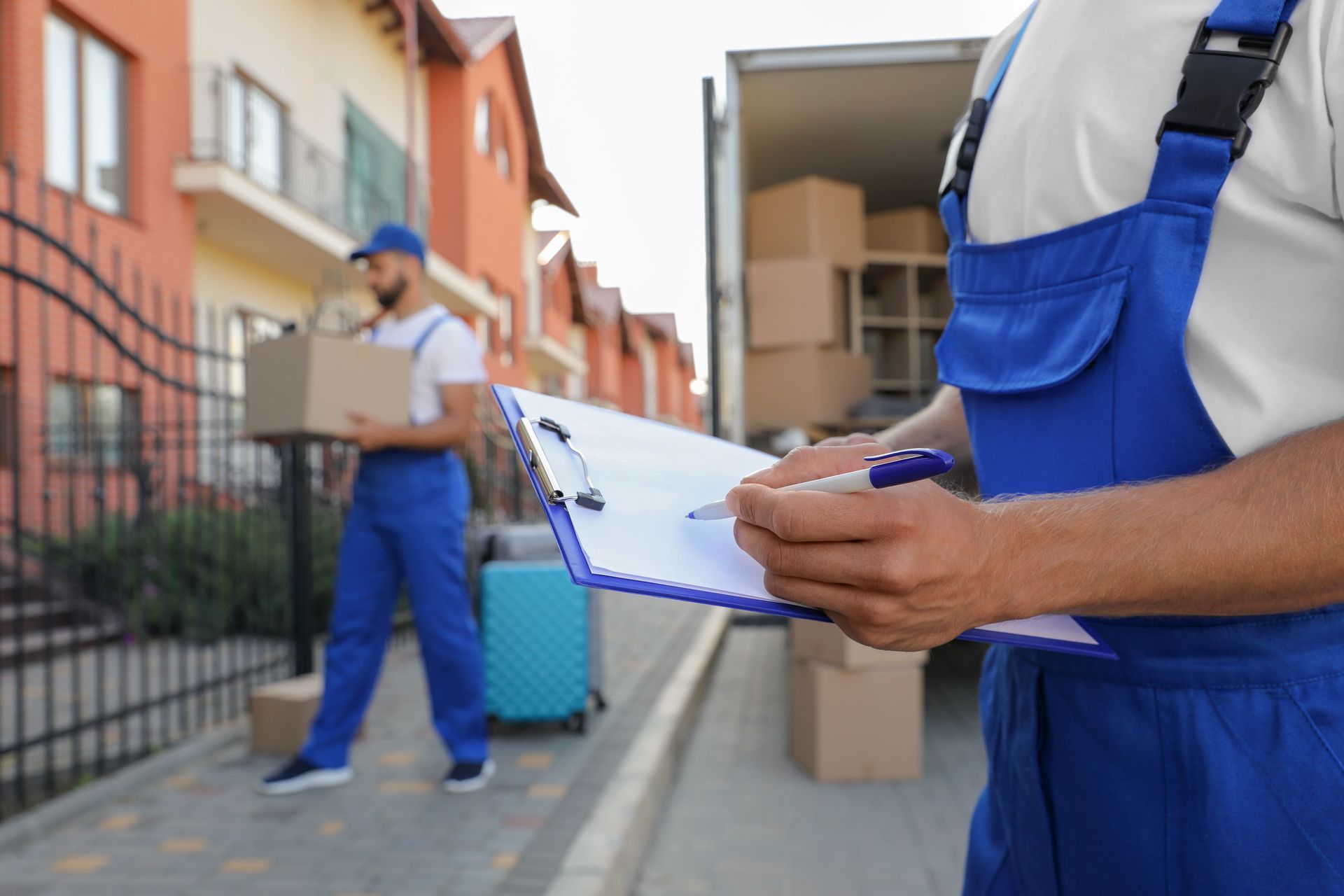 Movers loading boxes into a truck, one writing on a clipboard outdoors.