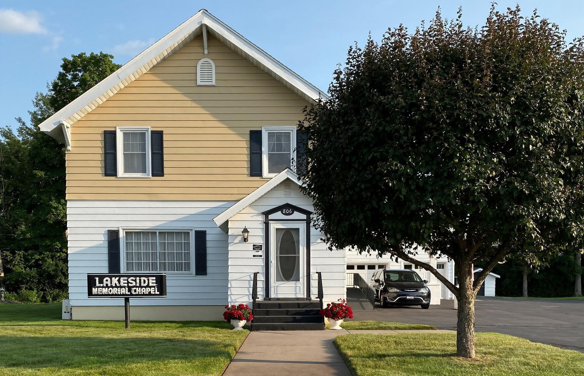Two-story house with tan and white siding, black shutters, and a dark tree in the front yard. A car is parked in the garage.