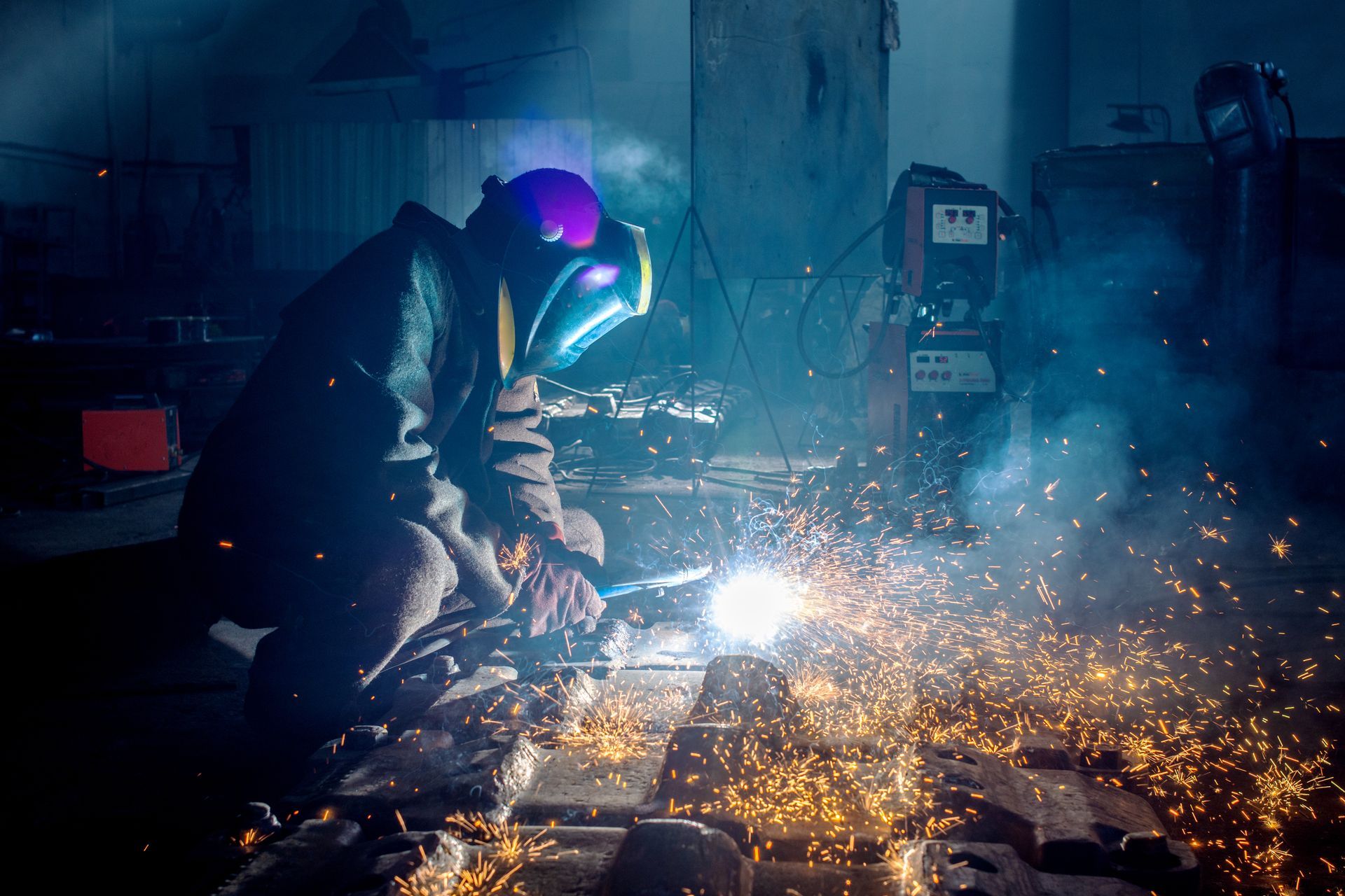 Side view of a steel fabricator in protective helmet welding metal detail with sparks at a factory.