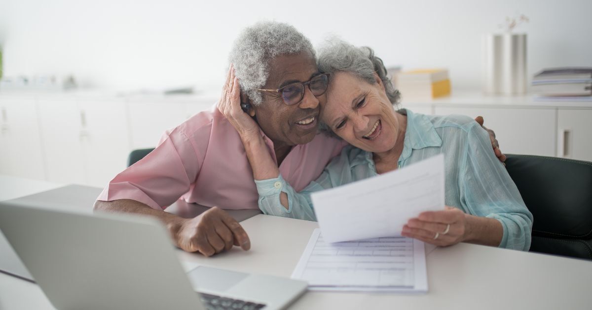 Elderly couple smiling, reading a document together at a table with a laptop. One has their arm around the other.