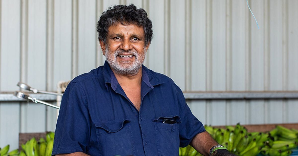 Smiling man in blue shirt, possibly a farmer, standing in front of a banana display.