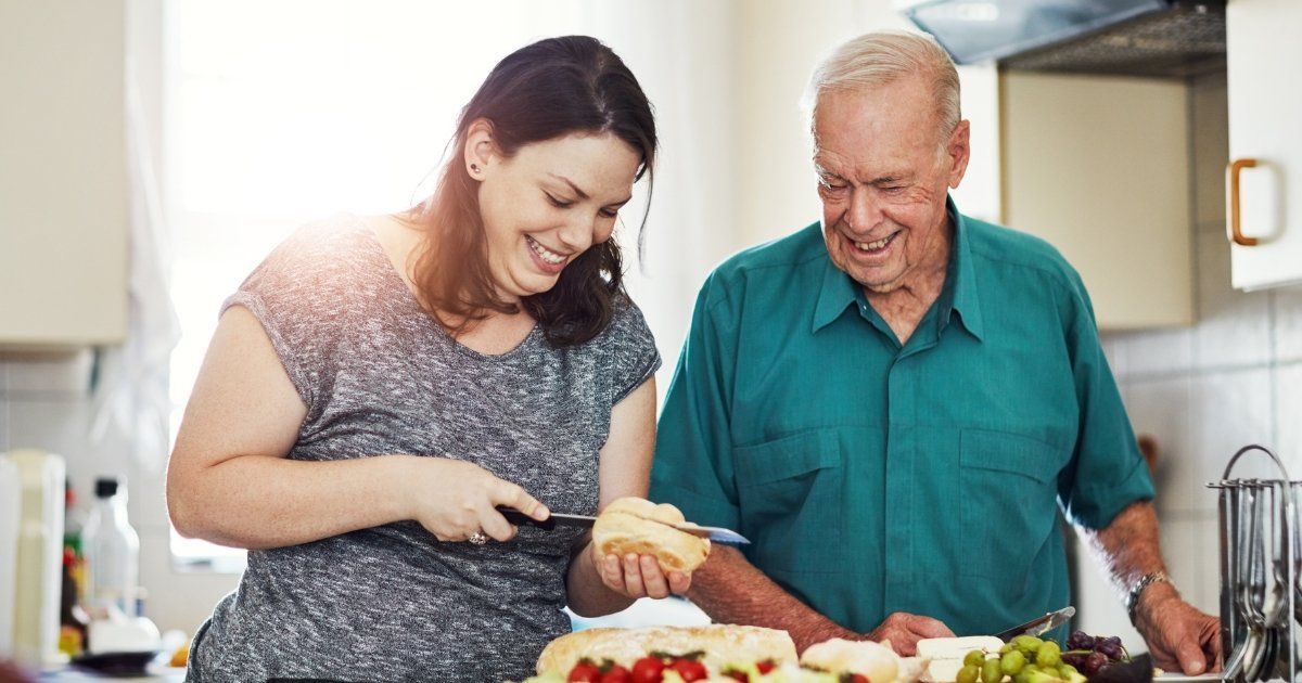 Woman and older man in a kitchen, smiling while the woman cuts a cake.
