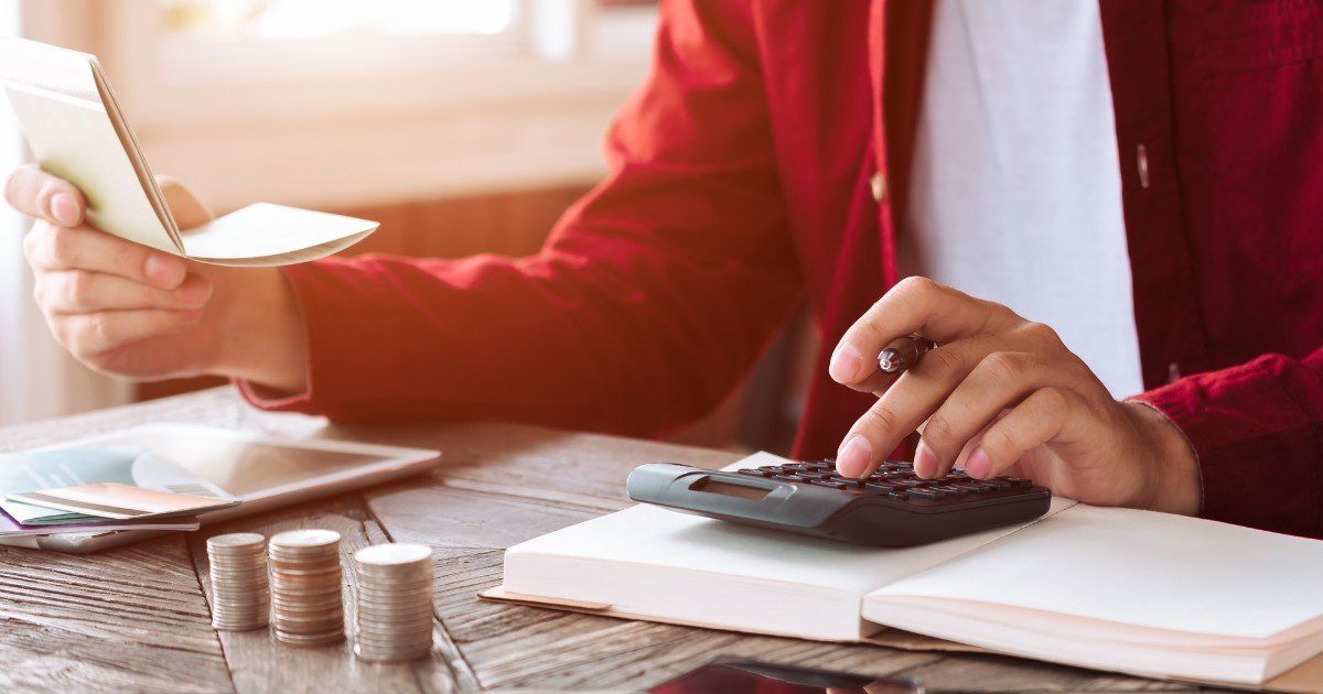 Person in a red jacket using a calculator with a notepad, coins, and a tablet on the desk, possibly budgeting.