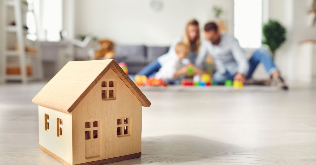 Wooden house model in focus; family playing with colorful blocks in background.