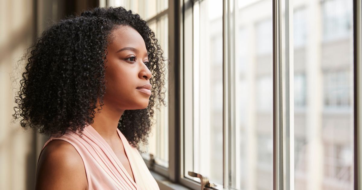 Woman with curly dark hair gazes thoughtfully out a window in a building, wearing a light pink top.