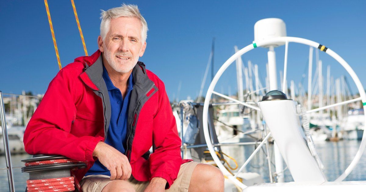 Man with gray hair wearing a red jacket, seated on a sailboat. The background shows boats docked at a marina.