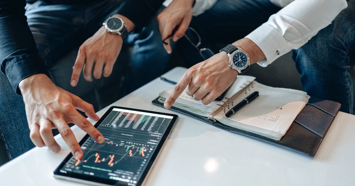 Two men pointing at a tablet displaying stock charts, seated at a table with a notebook and pen. Both men are wearing watches.