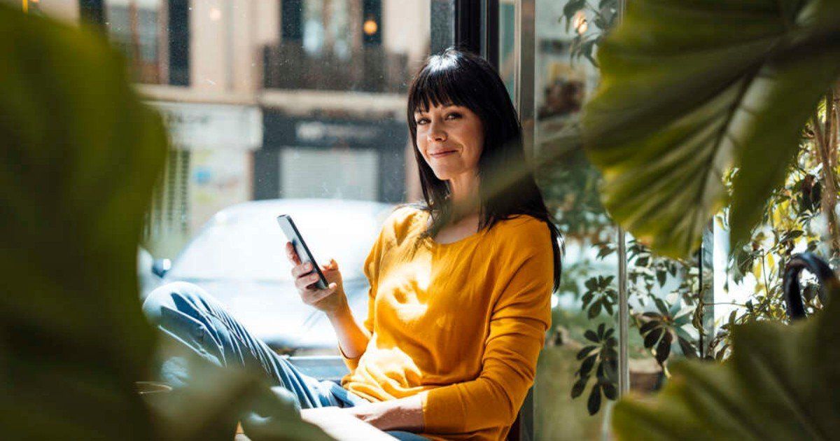 Woman with dark hair in a yellow sweater sits by a window, holding a phone, looking at the camera.