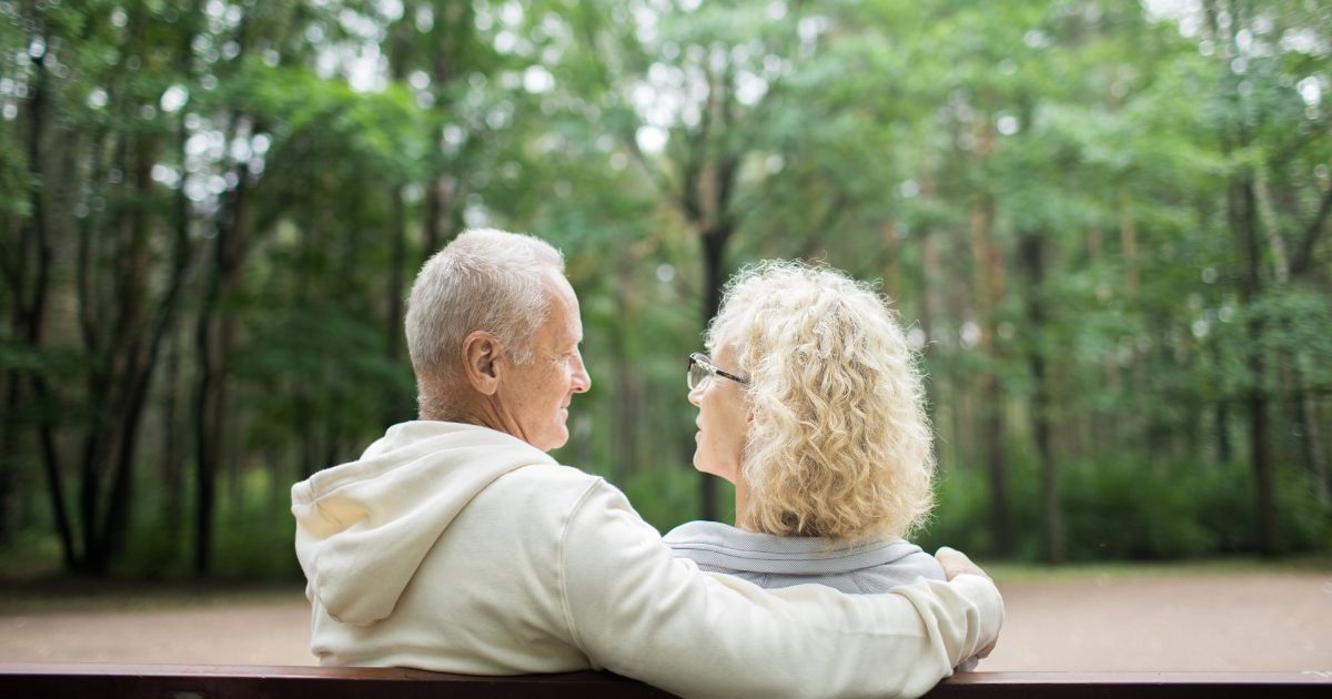 Older couple with arms around each other, sitting on a bench in a park. They are looking at each other lovingly.