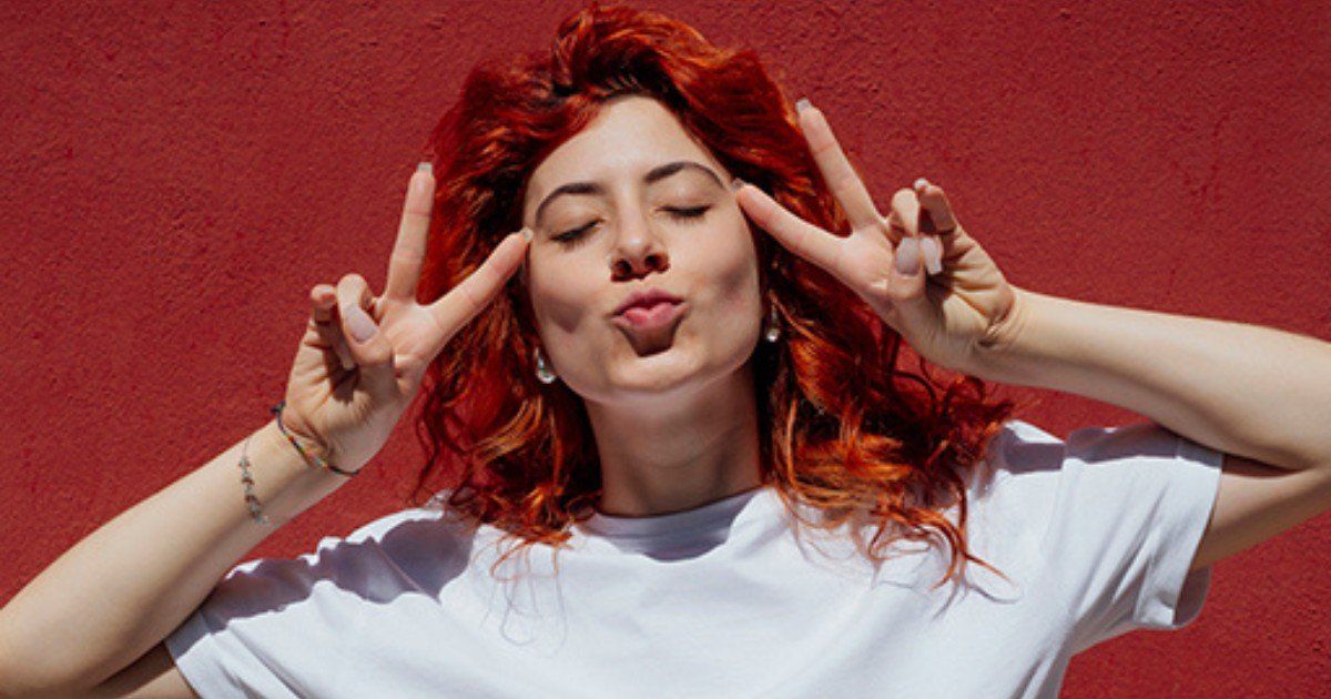 Woman with red hair making a peace sign with both hands near her eyes and blowing a kiss, against a red wall.