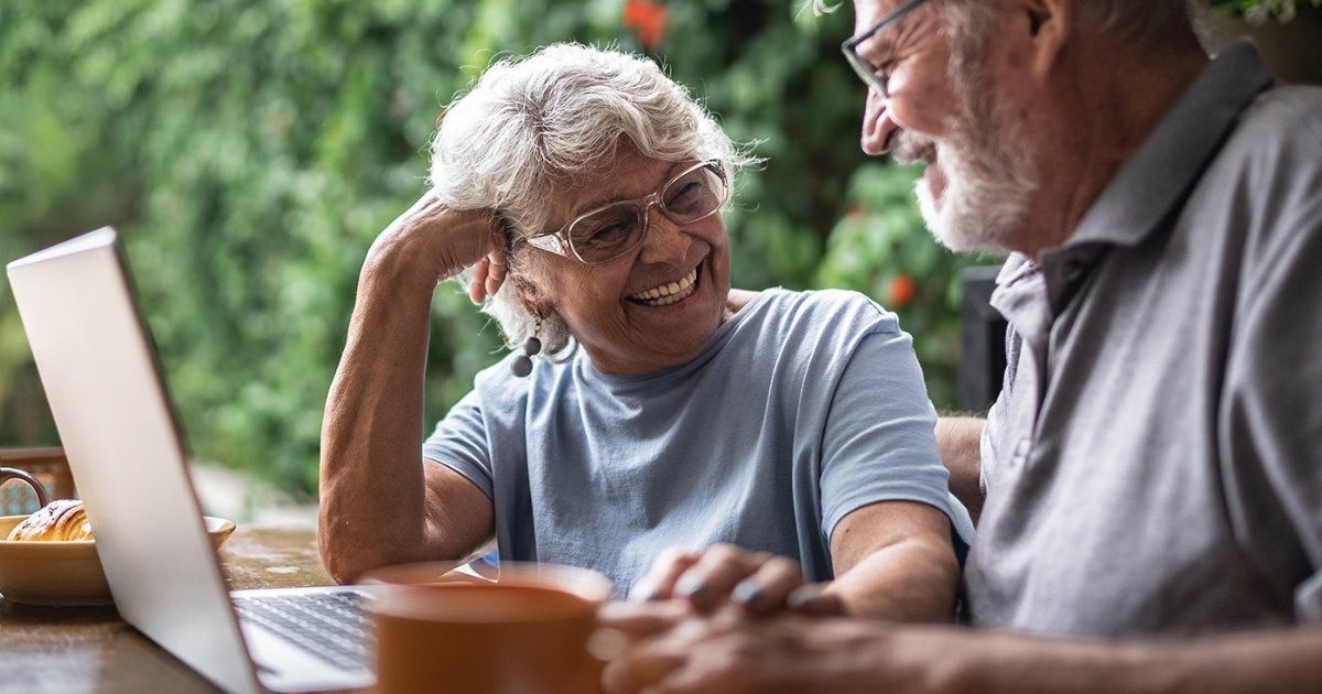 Elderly couple smiles together while looking at a laptop outdoors, woman resting her head on her hand, man's arm on hers.