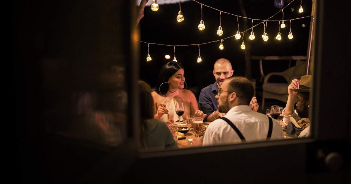 People at a table, illuminated by string lights, are enjoying an outdoor dinner party at night.