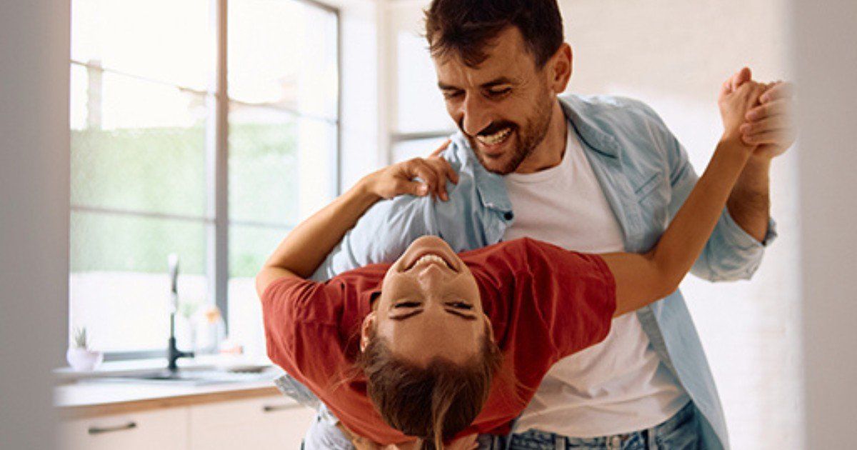 Man and woman laughing while dancing in a kitchen. The woman is leaning back, held by the man. Bright, natural light.