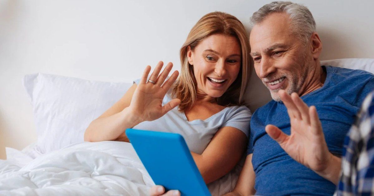 A smiling senior couple waves at a tablet, appearing to video chat from a bed. The woman wears a gray shirt, the man a blue shirt.