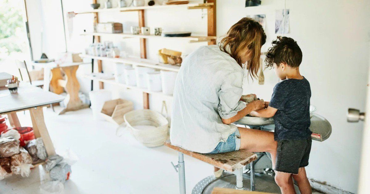 Woman and child working together at a pottery wheel in a bright workshop. The child is helping the woman shape the clay.