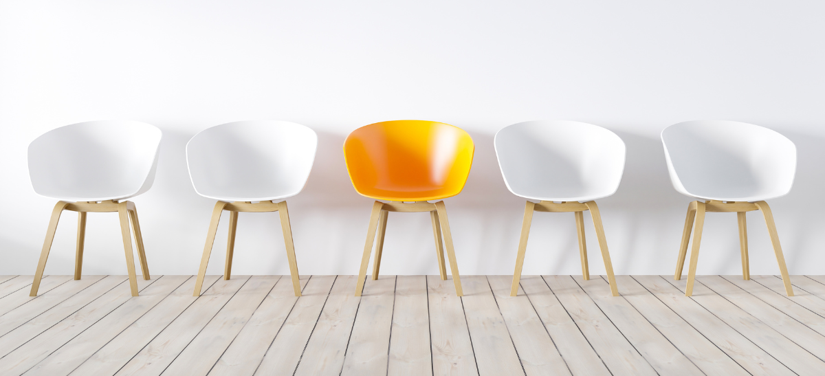 A single orange chair stands in the center of a row of four white chairs against a plain white wall.