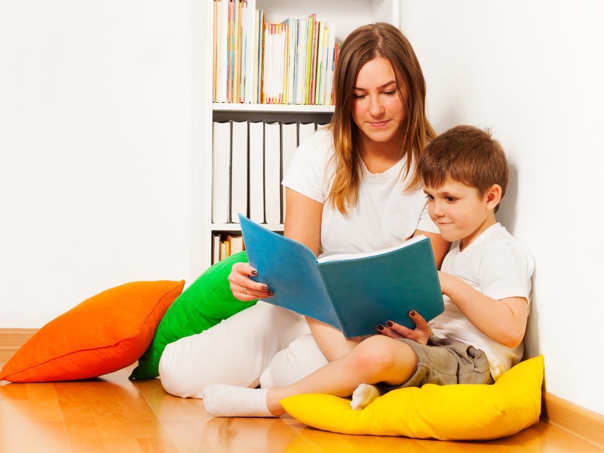 A person reads a blue book to a child while sitting on the floor near a bookshelf with colorful pillows.