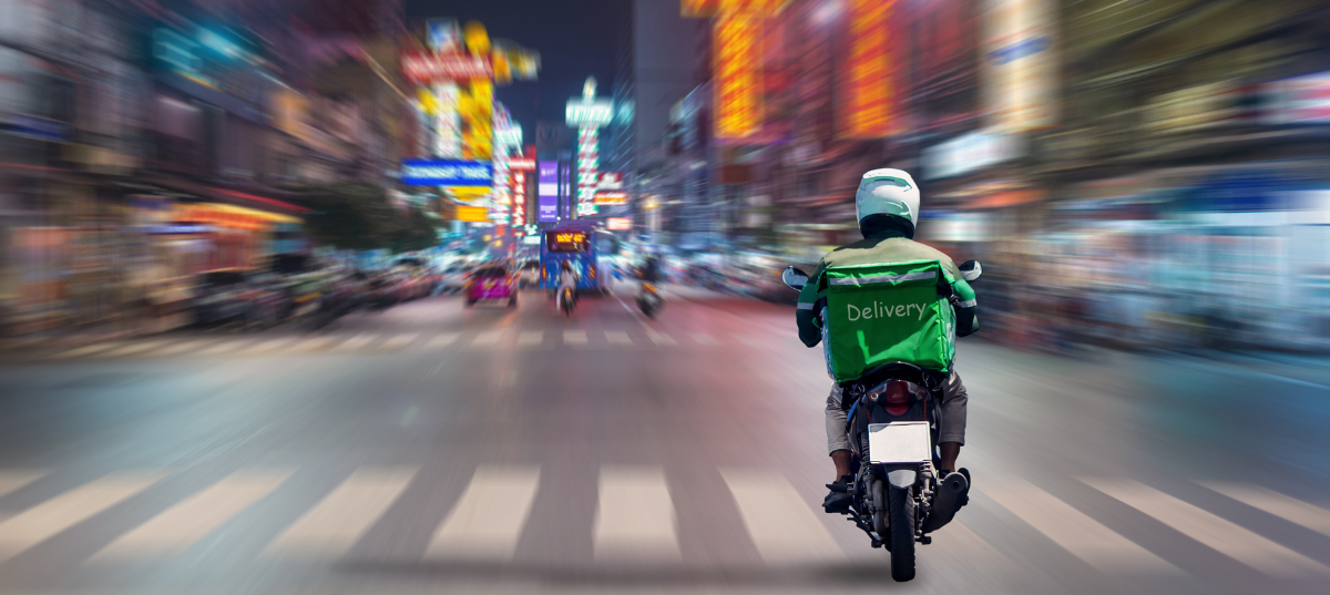 A delivery driver on a motorcycle drives across a crosswalk on a busy, motion-blurred city street at night.