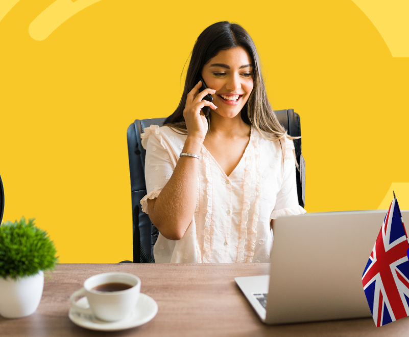 A person smiles while talking on a phone at a desk with a laptop, a coffee cup, a small plant, and a UK flag.