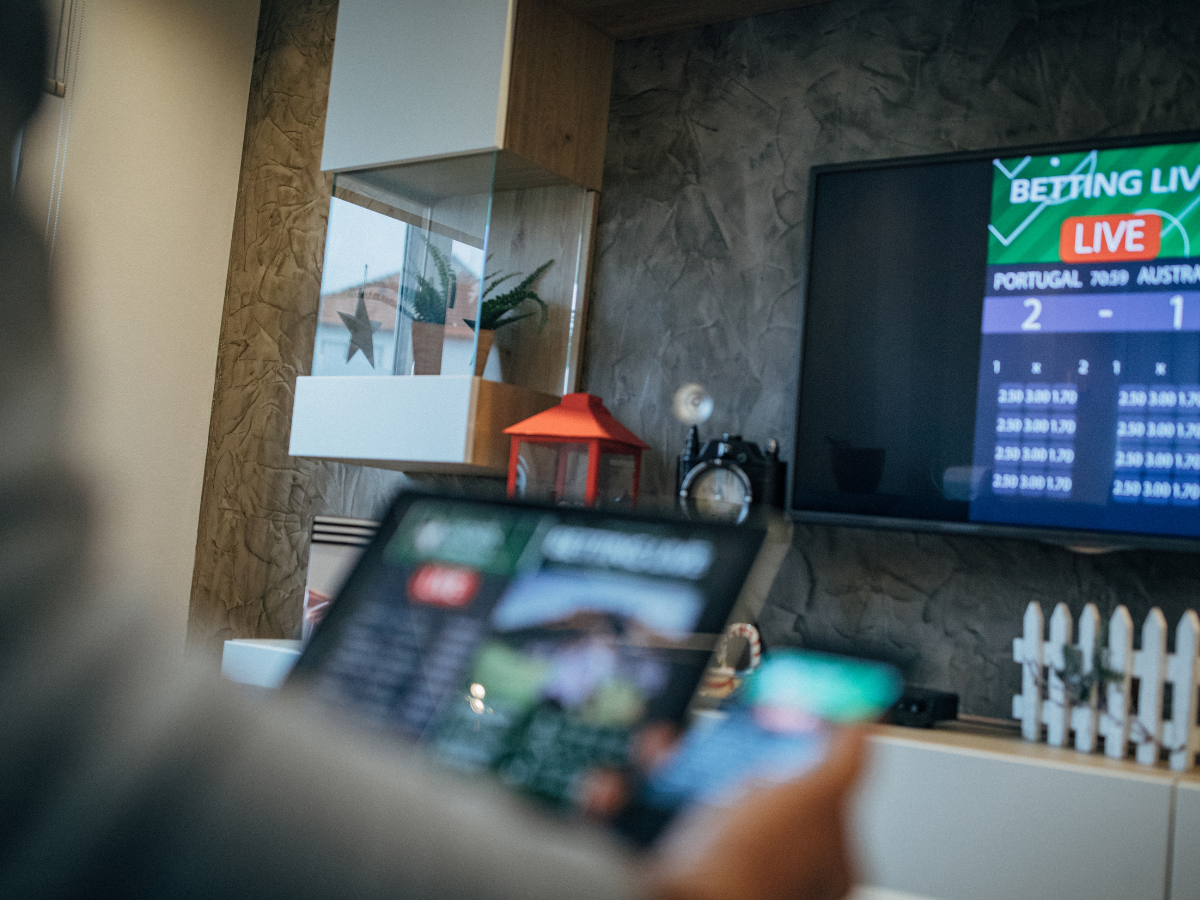 A person holds a tablet and phone while watching a live sports betting score board on a television screen.