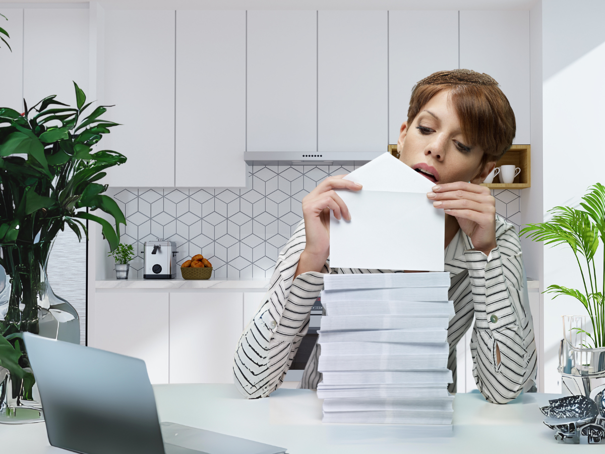 A person in a striped shirt opening a white envelope while seated at a table with a tall stack of mail in a modern kitchen.