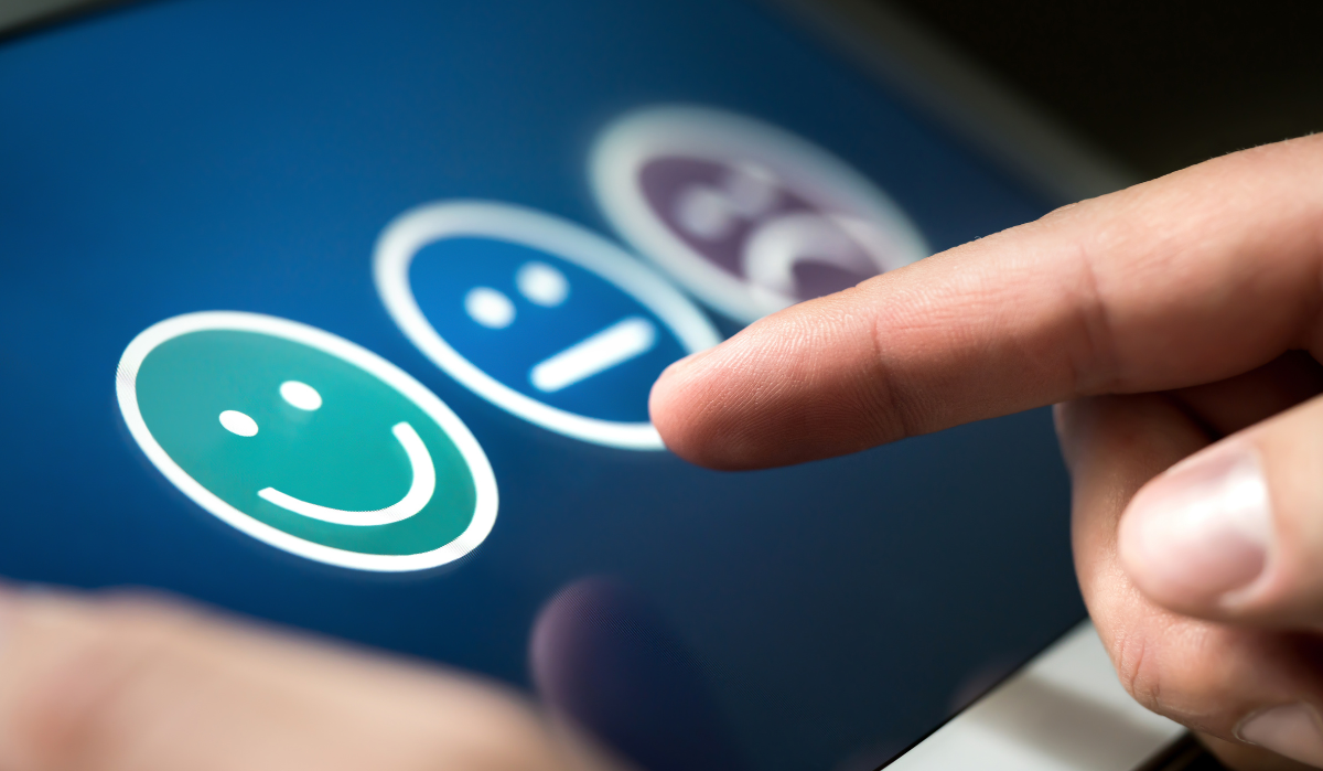 A hand touches a screen displaying green, blue, and purple rating icons with happy, neutral, and sad expressions.