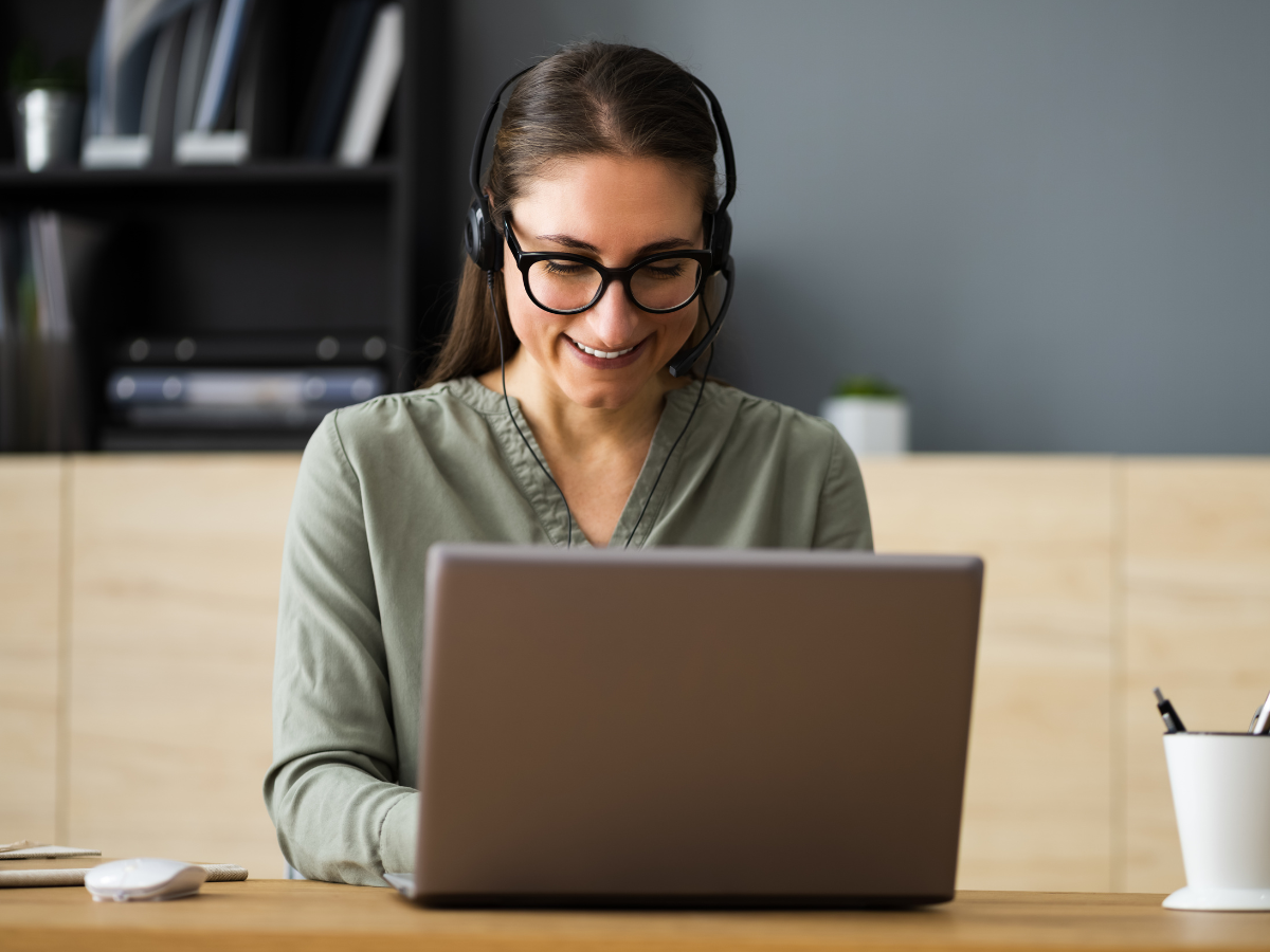 A person wearing a headset and glasses smiles while working on a laptop at a desk in an office setting.