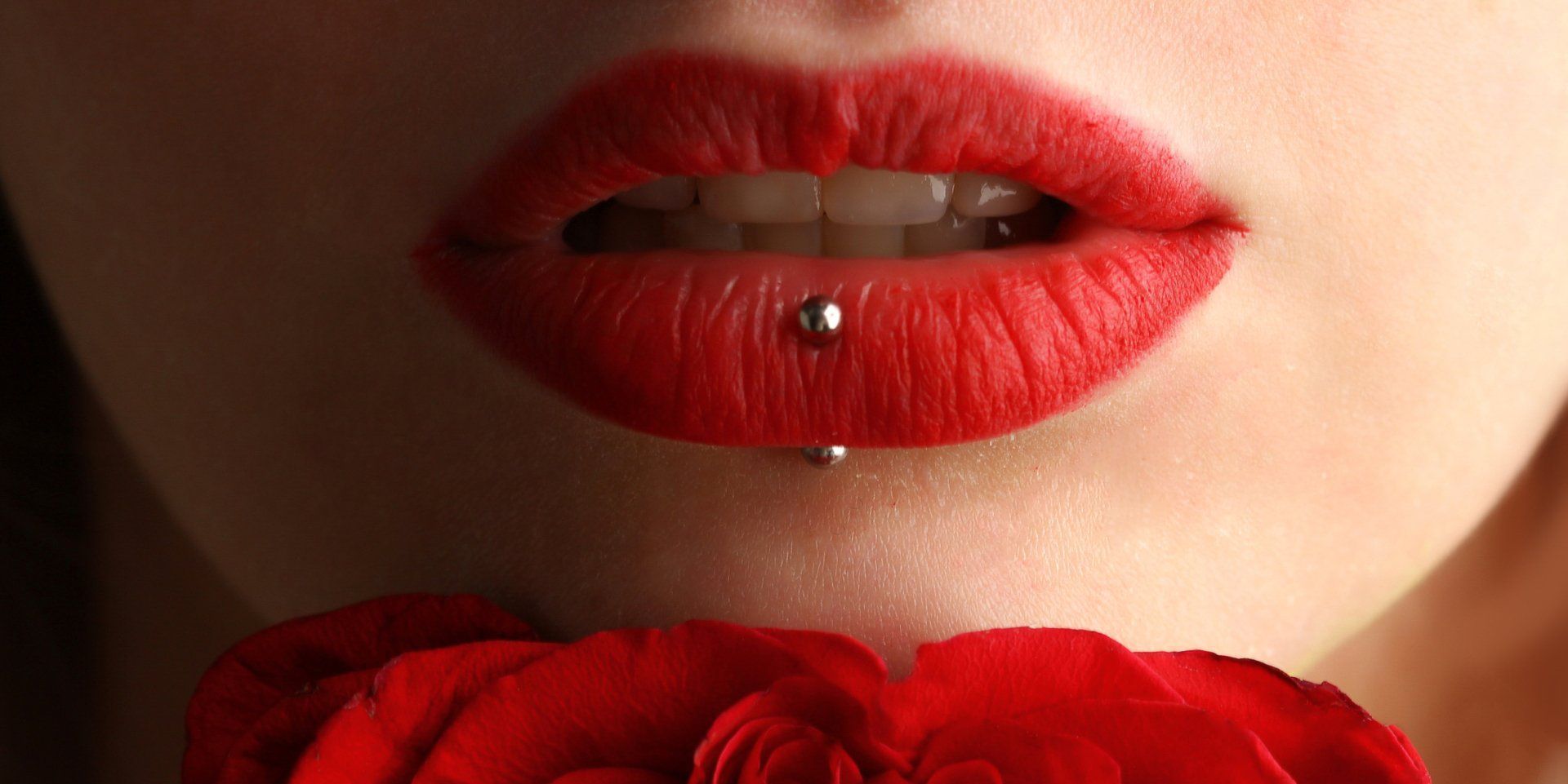 Close-up of bright red lips with a lower lip piercing, held just above a red rose.