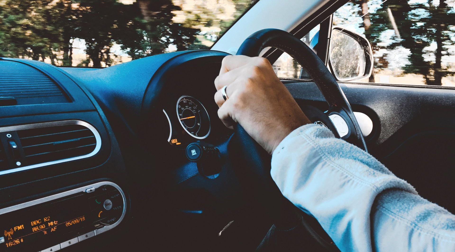 A person drives a car with their hand on the steering wheel while looking out the window at a blurry, sunlit landscape.