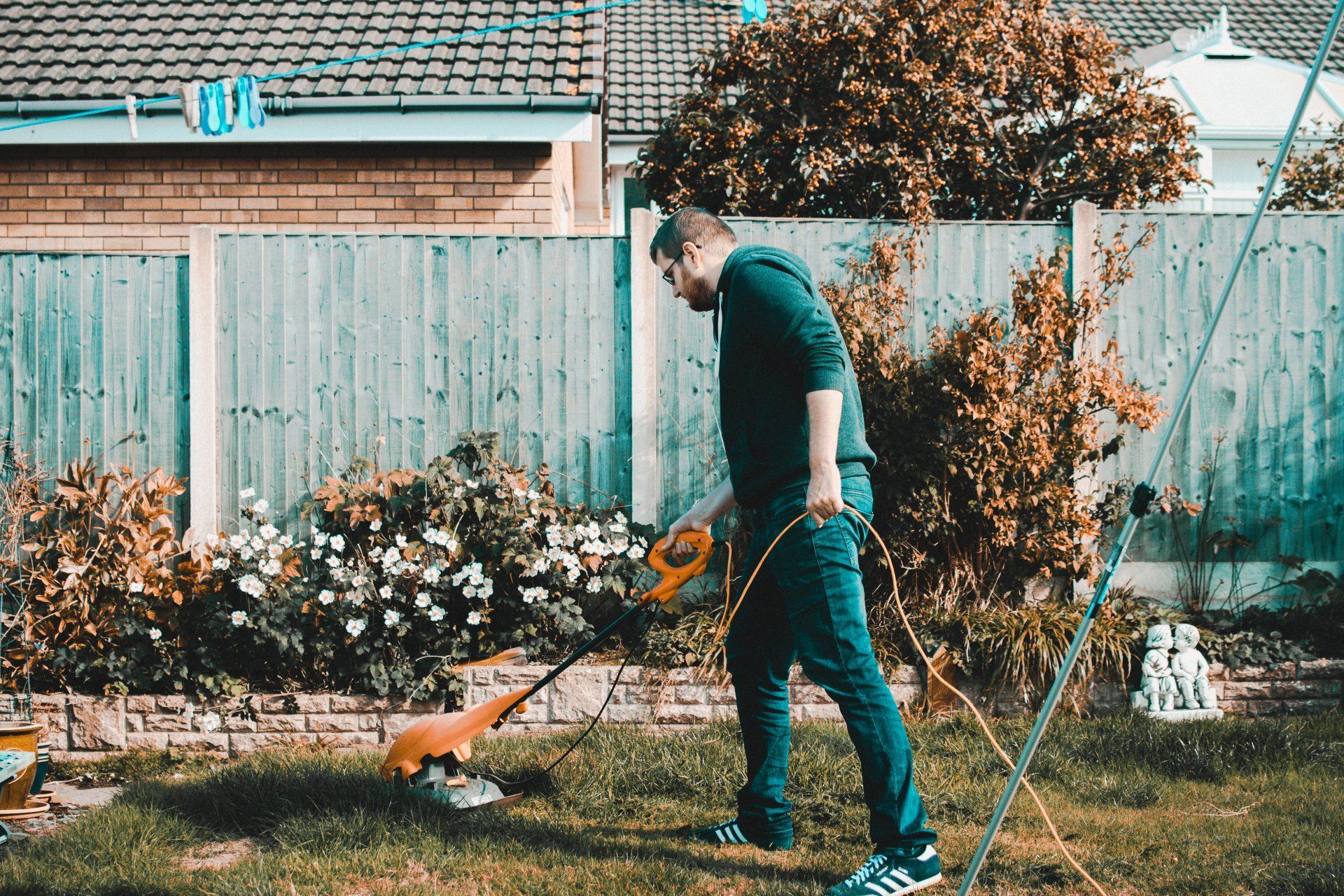 A person in a green hoodie uses an orange string trimmer to clear grass in a backyard.