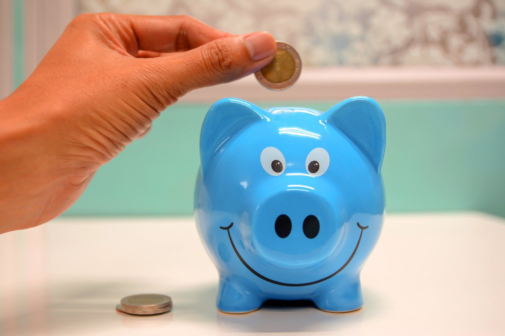 A person's hand deposits a coin into a bright blue, smiling piggy bank sitting on a white surface.